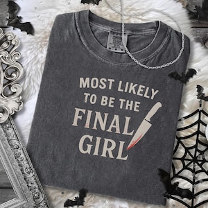 May include: Dark gray t-shirt with the text "MOST LIKELY TO BE THE FINAL GIRL" and a graphic of a bloody knife. The shirt is surrounded by Halloween decorations, including bats, a spider web, and a candle.