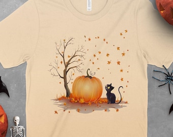May include: Beige t-shirt featuring a Halloween-themed graphic. The design includes a large orange pumpkin, a black cat, a leafless tree, and falling leaves. The shirt is surrounded by Halloween decorations, including pumpkins, bats, and spiders.