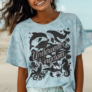 以下が含まれることがあります： 水色のタイダイ染めのTシャツに、黒い海の生き物のグラフィックと「Underwater Kingdom」の文字がプリントされています。