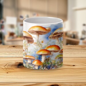 Può includere: Tazza in ceramica bianca con un'illustrazione ad acquerello di funghi con cappelli arancioni e gambi bianchi. Il design include uno sfondo di cielo blu e fogliame verde, creando un tema ispirato alla natura.