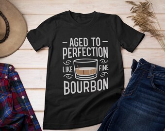 May include: A black short-sleeved t-shirt featuring white text that reads AGED TO PERFECTION LIKE FINE BOURBON, accompanied by a graphic illustration of a glass containing a brown liquid and a single ice cube centered on the chest.