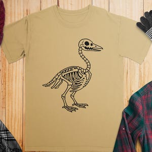 May include: A tan t-shirt featuring a black graphic of a bird skeleton. The skeletal design is detailed, showing the rib cage, spine, and skull. The shirt is displayed on a wooden surface with other clothing items.