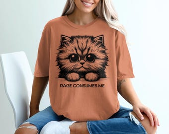 May include: A burnt orange t-shirt featuring a black and white illustration of a fluffy cat with large eyes. The text "RAGE CONSUMES ME" is printed below the cat image. The shirt is a casual wear item.