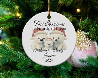 Puede incluir: Adorno navideño redondo y blanco con una familia de osos polares con gorros de Papá Noel. El texto dice "First Christmas as Family of three", con "Mommy Daddy Jennifer 2024" debajo. El adorno cuelga de un árbol de Navidad.