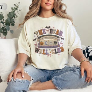 May include: An ivory short-sleeve t-shirt featuring a graphic print with the text "THE DEAD WALL CLUB" and illustrations of a pumpkin, moon, ghost, and black cat arranged like mahjong tiles, surrounded by bats, stars, and small pink bows.