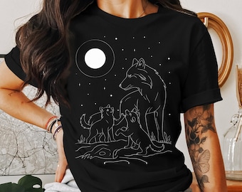 May include: A black short-sleeved t-shirt featuring a white line art graphic of a large wolf standing over two smaller wolf pups. The scene includes a full moon and scattered stars in the background, set against a dark, solid-colored fabric.