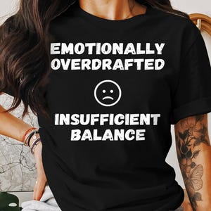 Könnte beinhalten: Schwarzes T-Shirt mit weißem Text, der "Emotionally Overdrafted Insufficient Balance" lautet, mit einem weißen Emoji mit traurigem Gesicht.