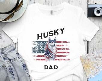 May include: White t-shirt with a distressed American flag design and a husky dog wearing a red, white, and blue bandana. The text "HUSKY DAD" is printed on the shirt.