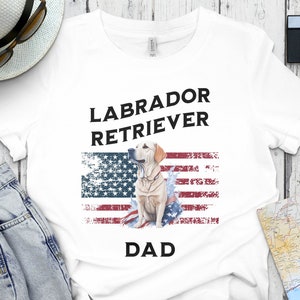 Peut inclure: T-shirt blanc avec un motif de drapeau américain usé et le texte "Labrador Retriever Dad". Un labrador retriever jaune se tient devant le drapeau.