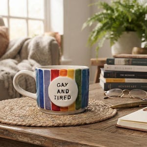 Pode incluir: Uma caneca de cerâmica com listras verticais de arco-íris em azul, roxo, vermelho, laranja, amarelo e verde. Um emblema circular branco em relevo na frente exibe o texto preto em relevo GAY AND TIRED. A caneca tem uma alça curva e base larga.