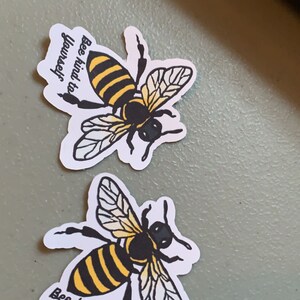 Könnte beinhalten: Zwei Bienen-Sticker mit dem Text "Bee kind to yourself" auf weißem Hintergrund. Die Bienen sind schwarz und gelb mit weißen Flügeln.