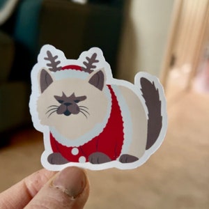 May include: A sticker featuring a cartoon cat wearing a red Santa suit with white trim and reindeer antlers. The cat has a grumpy expression and a brown tail. The sticker is on a white background.