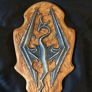 May include: A wooden plaque with a silver dragon symbol, the dragon is in a curled position with its wings spread out. The plaque has a wood grain pattern.