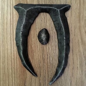 May include: A dark gray, 3D-printed emblem featuring a stylized letter "U" shape with a central oval. The emblem has a textured surface and is set against a wood grain background. The design is reminiscent of a fantasy game logo.