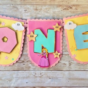 May include: A pink, yellow and blue banner with the word "ONE" in large letters. The banner is decorated with stars, clouds and a small figure of Princess Peach from the Super Mario Bros. video game series.