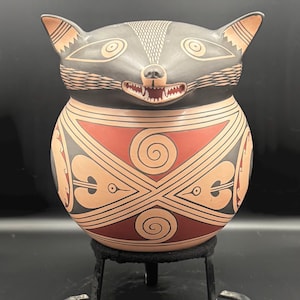 Può includere: Un vaso in ceramica a forma di procione con testa e orecchie nere e corpo rosso e beige. Il procione ha lineamenti dettagliati ed è appoggiato su un supporto in metallo nero. Il corpo presenta disegni geometrici.