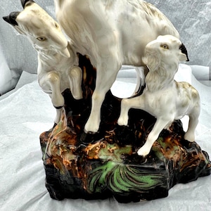 Op de afbeelding: Een keramische sculptuur van een gezin van geiten. De sculptuur toont een grote geit met twee kleinere geiten. De geiten zijn wit met zwarte accenten en staan op een voetstuk met bruine, groene en zwarte kleuren.