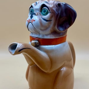 Könnte beinhalten: Keramik-Teekanne in Hundeform, braun mit weißer Brust und rotem Halsband. Das Gesicht des Hundes hat grüne Augen und schwarze Details. Der Ausguss bildet die ausgestreckte Pfote des Hundes.