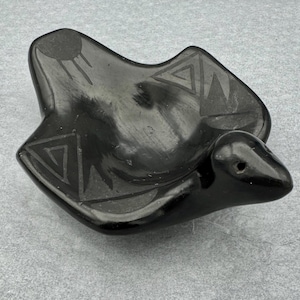 May include: A black ceramic bird-shaped bowl with geometric designs. The bowl features a sun and triangle patterns. The bird has a small hole on its beak. This decorative bowl is suitable for holding small items.