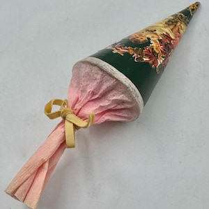 May include: A conical gift container featuring a dark green exterior with a floral and leaf print. The wide opening is filled with light pink crinkled tissue paper, which is gathered and secured with a small, pale yellow ribbon tied in a bow.