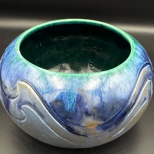 May include: A round ceramic bowl with a glossy finish. The bowl features a gradient of colors, transitioning from a deep teal interior to shades of blue and silver on the exterior. Decorative wave patterns adorn the sides.