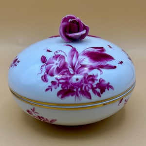May include: A white porcelain trinket box with a lid, adorned with a floral design in shades of purple. The lid is topped with a rose-shaped handle. The box has a gold trim around the base and lid.
