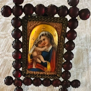 May include: A small framed image of the Virgin Mary holding a baby Jesus. The frame is made of dark red beads and has a gold-coloured border. The image is on a white background.