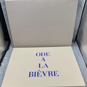以下が含まれることがあります： 明るいタン色の表紙と青い文字で「ODE A LA BIÈVRE」と書かれた開いた本。本は、「Louise Bourgeois」のエンボス加工が施された明るいタン色のスリップケースに入っています。本は明るい灰色の表面にあります。