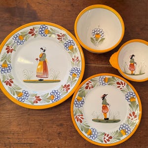 Könnte beinhalten: Ein Set aus Keramikgeschirr mit einem Volkskunst-Design. Das Set umfasst Teller und Schalen mit weißem Hintergrund, gelben Rändern und floralen und figürlichen Illustrationen. Die Teller und Schalen sind auf einer Holzoberfläche angeordnet.