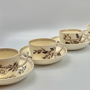 Może przedstawiać: Zestaw trzech kremowych filiżanek ceramicznych z pasującymi spodkami, ozdobionych brązowym botanicznym wzorem kwiatowym oraz powtarzającym się geometrycznym obrzeżem na górnej krawędzi filiżanek i zewnętrznym brzegu spodków.
