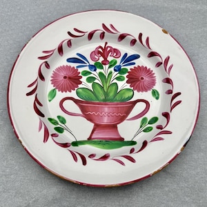 May include: A white ceramic plate with a hand-painted floral design. The plate features a central arrangement of flowers in a red vase, surrounded by pink and blue flowers, green leaves, and a red border. The plate's diameter is approximately 10 inches.