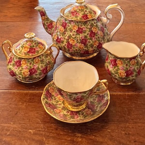 Könnte beinhalten: Ein florales Teeservice mit Teekanne, Zuckerdose, Sahnekännchen, Tasse und Untertasse. Das Set hat einen grünen Hintergrund mit roten und gelben Rosen und goldenen Verzierungen. Die Teekanne hat einen geschwungenen Griff und einen Deckel mit goldener Spitze.