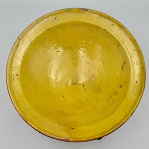 May include: A circular ceramic bowl with a vibrant yellow glazed interior surface. The bowl features a wide, flat rim and a smooth, glossy finish with subtle natural variations and small dark speckles visible throughout the yellow glaze.