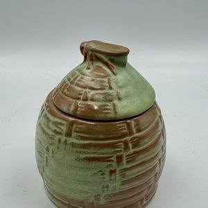 Może przedstawiać: Ceramiczny słoik z pokrywką w kształcie tradycyjnego ula. Przedmiot posiada teksturowany wzór w stylu plecionki w naprzemiennych odcieniach ziemistego brązu i delikatnej szałwiowej zieleni, z małą rzeźbioną figurką pszczoły na pokrywce.