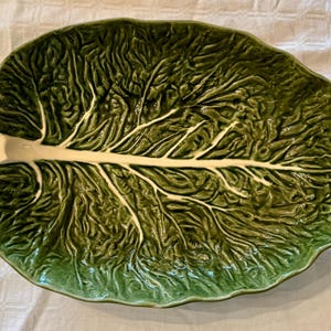 Op de afbeelding: Een groene keramische serveerschaal in de vorm van een koolblad. De schaal heeft een textuuroppervlak met gedetailleerde nerven en een crèmekleurige steel. Het ontwerp is een realistische weergave van een koolblad.