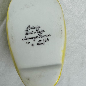 Pu&ograve; includere: Un piatto in ceramica bianca con bordo giallo. Il piatto presenta il testo "Artoria Peint Main Limoges France" e il simbolo del copyright Disney. Il piatto &egrave; contrassegnato con il numero 164.