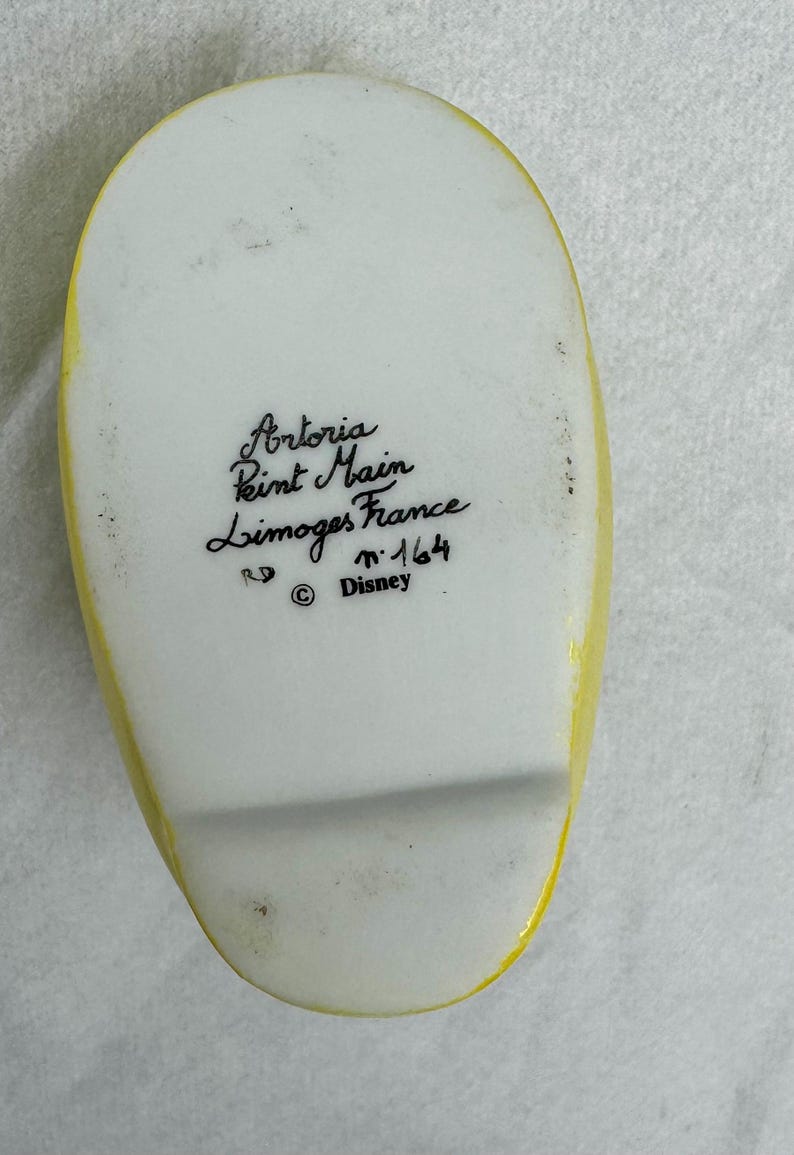 Pu&ograve; includere: Un piatto in ceramica bianca con bordo giallo. Il piatto presenta il testo "Artoria Peint Main Limoges France" e il simbolo del copyright Disney. Il piatto &egrave; contrassegnato con il numero 164.
