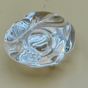 May include: A clear, swirled glass object with a central circular indentation. The transparent material reflects light, creating a rippled effect. The design is organic and abstract, suggesting a decorative piece or art object.
