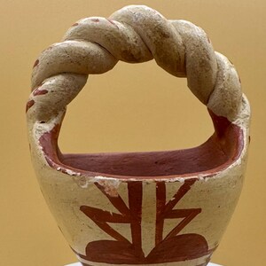 Puede incluir: Una pequeña cesta de cerámica pintada a mano con un asa trenzada. La cesta presenta un exterior de color crema con diseños geométricos rojos y marrones. El interior es de color marrón rojizo sólido. El asa es un diseño de cuerda retorcida con detalles rojos.