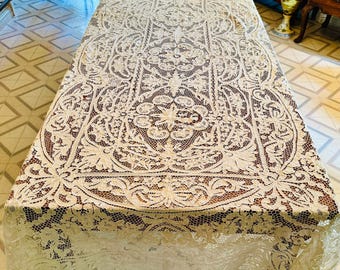 May include: A delicate white lace tablecloth with intricate floral patterns. The tablecloth is laid out on a wooden table.