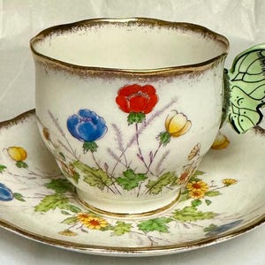 Op de afbeelding: Crèmekleurige theekop en schotel set met een gouden rand en een vlindervormige handgreep. De kop en schotel zijn versierd met kleurrijke bloemmotieven, waaronder rode, blauwe en gele bloemen en groene bladeren.