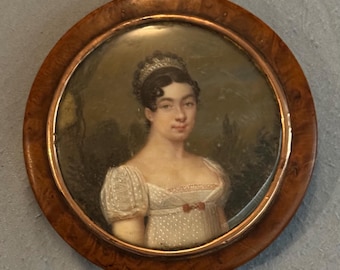 May include: A miniature portrait of a woman in a white dress with a low neckline and short sleeves. The portrait is set in a round, wooden frame with a dark finish. The woman has dark hair and is wearing a tiara.