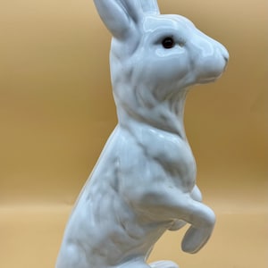 May include: A white ceramic rabbit figurine in a seated pose with its ears upright. The rabbit has red eyes and is on a light brown background. The figurine is a decorative item.