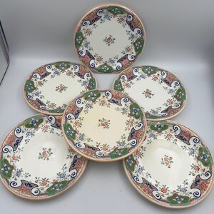 May include: A set of five round ceramic plates featuring a cream-coloured centre with a small floral motif. The wide rims are decorated with an intricate pattern of blue, green, pink, and orange floral and scroll designs on a white background.