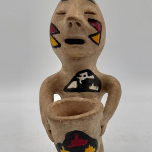 May include: A tan clay figurine of a kneeling person holding a small bowl. The figure features painted geometric designs in red, yellow, black, and white on the face, chest, and the bowl. The person has closed eyes and an open mouth expression.