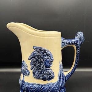 May include: A ceramic pitcher with a cream-colored body and blue accents. The pitcher features a raised relief of a Native American chief in profile, along with stylized trees and grass. The handle and base are blue, and the spout is cream-colored.