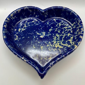 May include: A heart-shaped ceramic dish with a speckled blue and yellow pattern. The dish has a deep interior and a smooth, glossy finish. The edges are slightly raised, and the overall design is symmetrical.