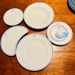 Op de afbeelding: Een set witte keramische borden met blauwe rand en het woord "Cafe" in blauwe schrift. De set bevat een groot dinerbord, een kleiner saladebord, een soepkom, een schoteltje en een halvemaanvormig bord.