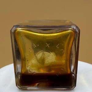 May include: A vintage, square glass bottle with a gold interior and a dark brown liquid. The bottle has a clear, faceted exterior and etched text inside the gold. The bottle is on a white surface. The bottle's contents are visible.