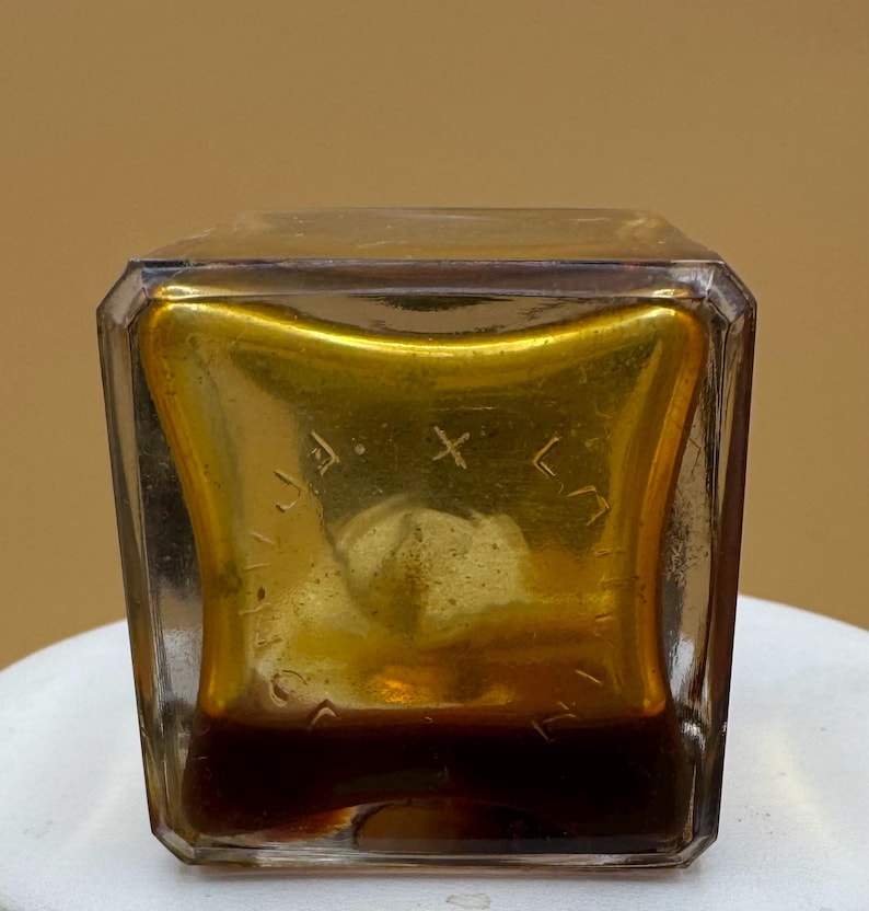 May include: A vintage, square glass bottle with a gold interior and a dark brown liquid. The bottle has a clear, faceted exterior and etched text inside the gold. The bottle is on a white surface. The bottle's contents are visible.